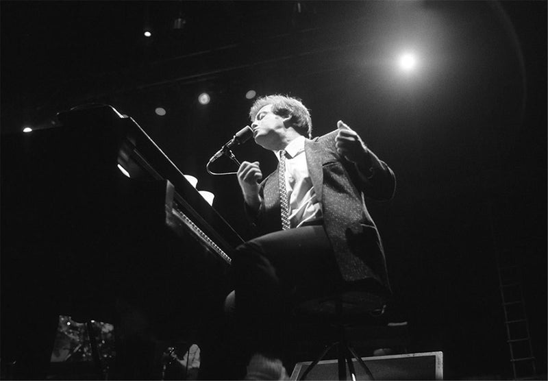 Billy Joel in sunglasses singing into the microphone from the piano, photographed by Charlyn Zlotnik.