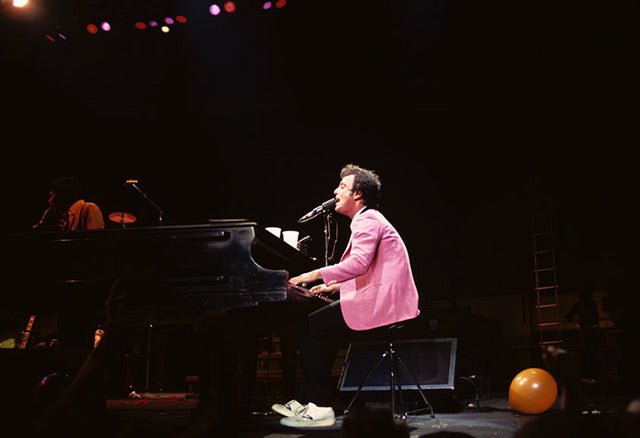 Billy Joel playing a grand piano in a pink jacket with an orange balloon on the stage floor, photographed by Charlyn Zlotnik.