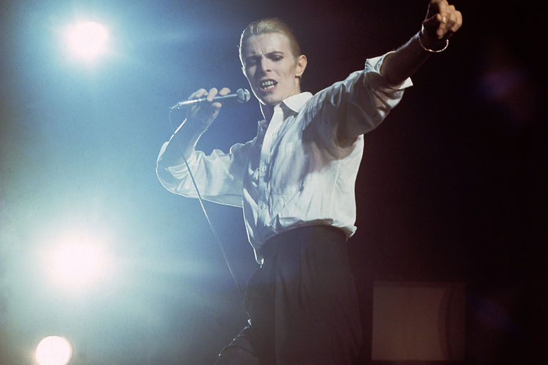 David Bowie performing with one arm raised against stage lights in 1976. Photograph by Gijsbert Hanekroot.