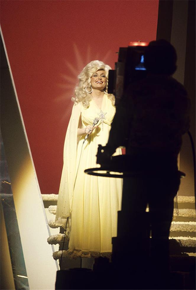 Dolly Parton on a television soundstage in a pale gown with a studio camera in silhouette, photographed by Charlyn Zlotnik.