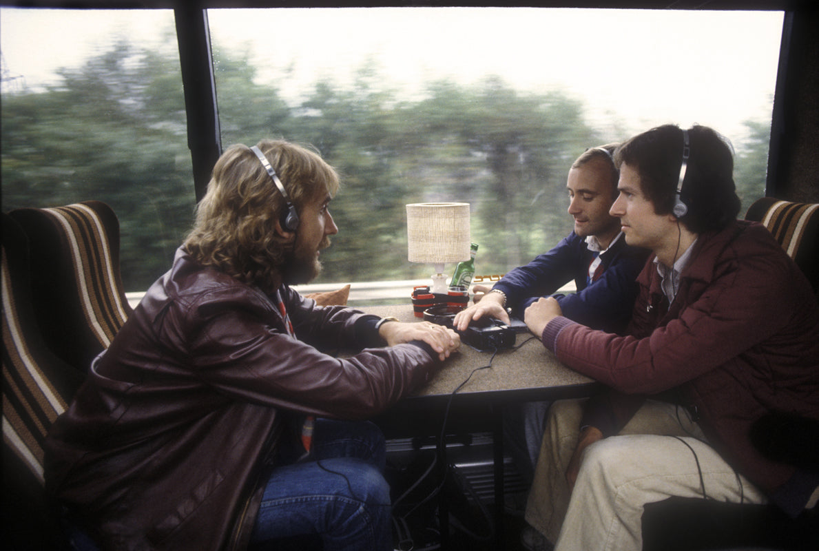Genesis, Phil Collins, Mike Rutherford and Tony Banks, sharing headphones around a table on the tour coach, photographed by Martyn Goddard.