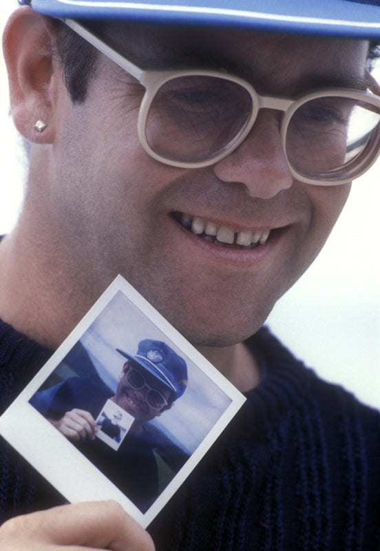 Elton John in a cap and oversized glasses holding a Polaroid of himself, photographed by Martyn Goddard, 1982.