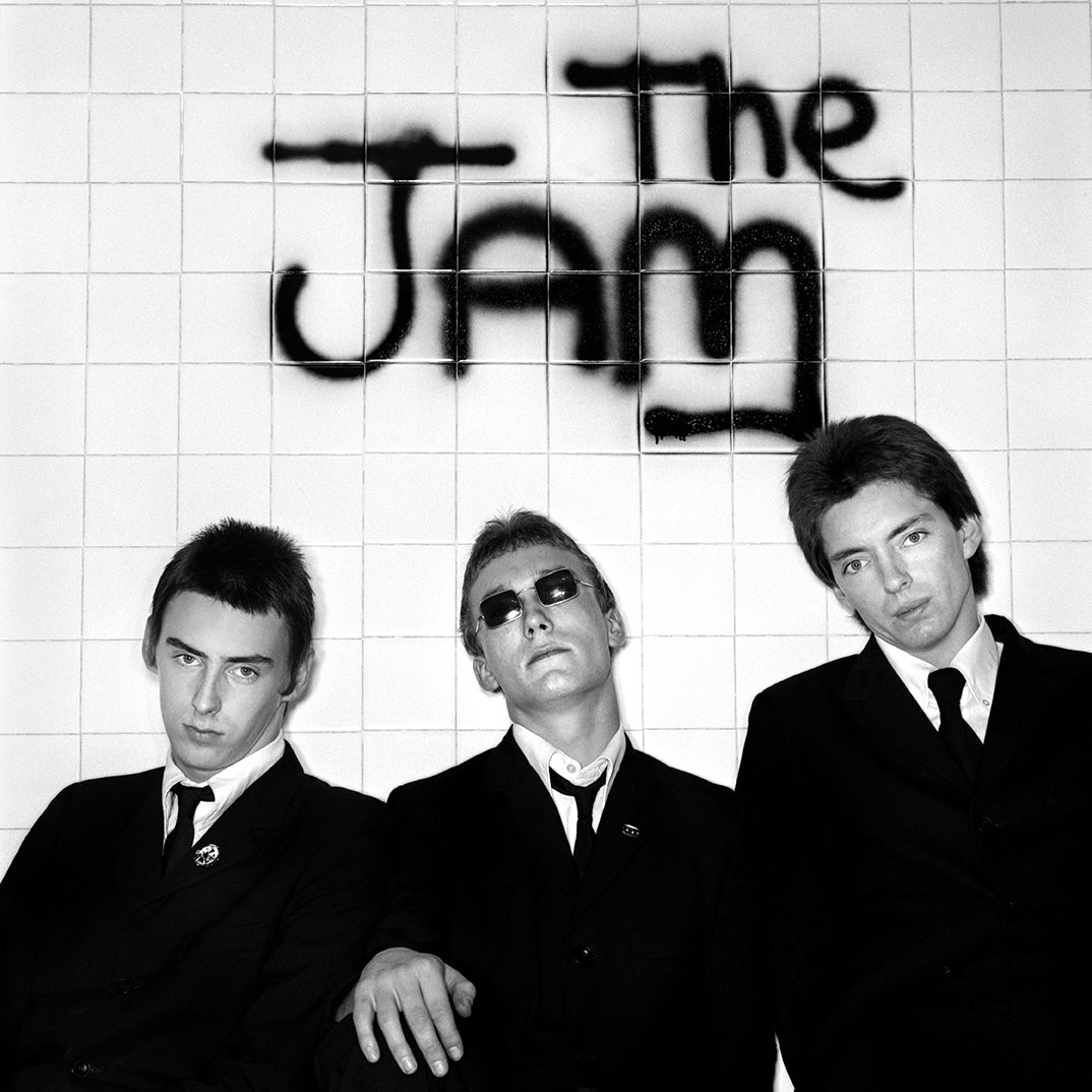 The Jam in dark suits against a white-tiled wall spray-painted with the band's name, In the City session, photographed by Martyn Goddard, 1977.