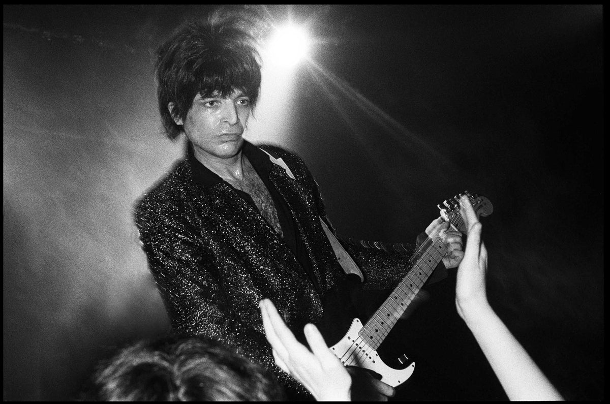 Alan Vega in a sparkling jacket playing guitar under a bright stage light as fans reach toward him, The Venue, London, 1982, photographed by David Corio.