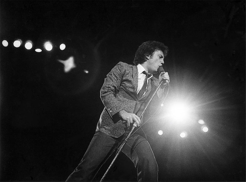 Billy Joel singing at the microphone stand under a spotlight, photographed by Charlyn Zlotnik.