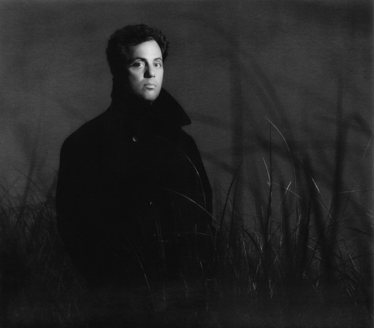 Billy Joel emerges from a dark backdrop in a restrained portrait, one hand raised toward his head. Photograph by Timothy White, East Hampton, New York, 1989.