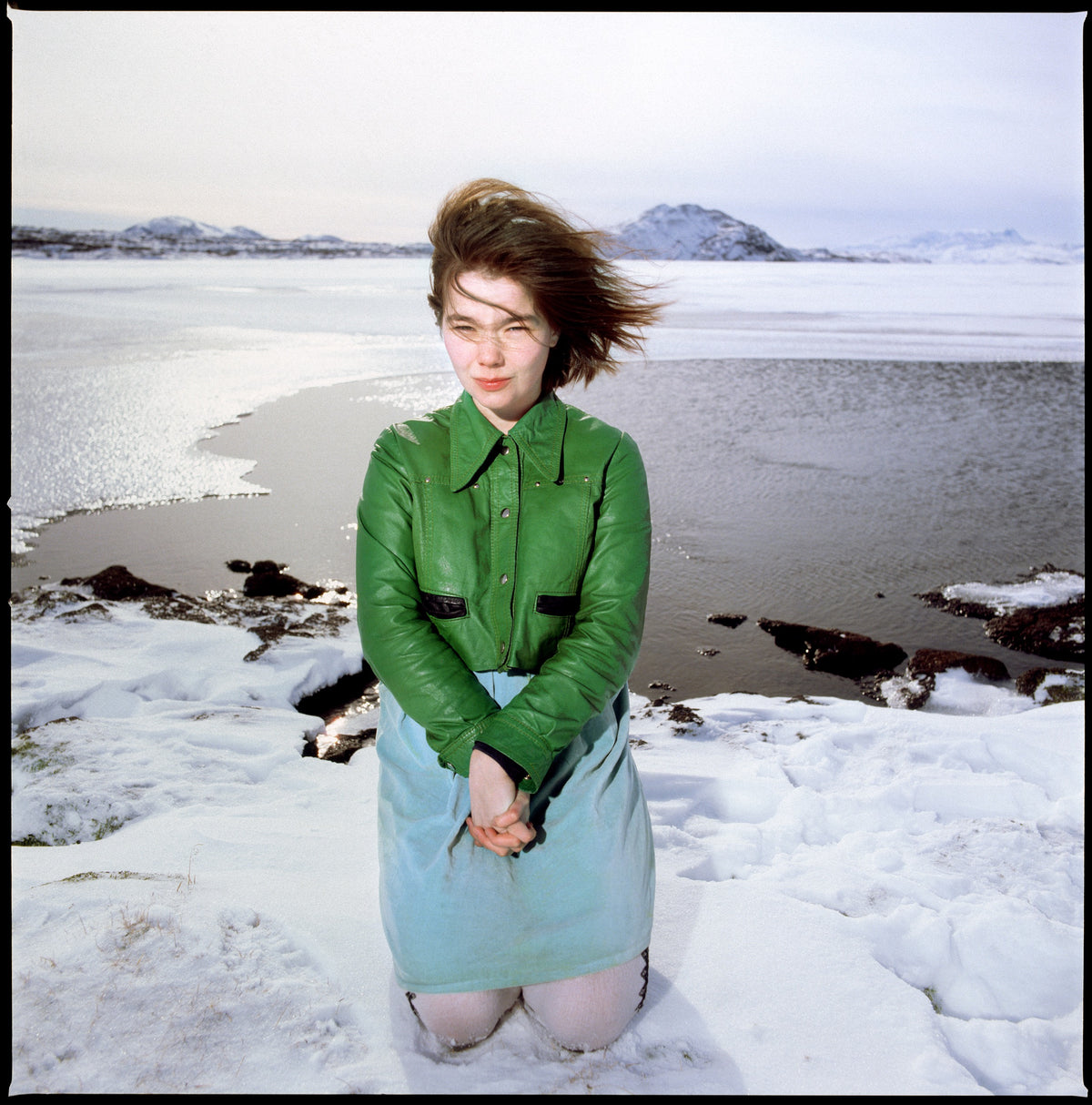 Wearing a green sweater, Björk kneels in a snowy coastal landscape with distant mountains behind her. Photograph by Timothy White, Reykjavík, Iceland, 1988.