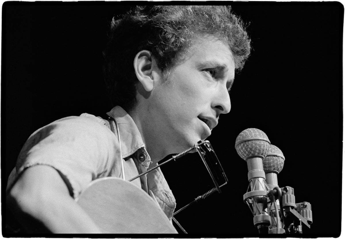 Bob Dylan singing into the microphone while playing guitar at the Newport Folk Festival in 1963, photographed by Rowland Scherman.