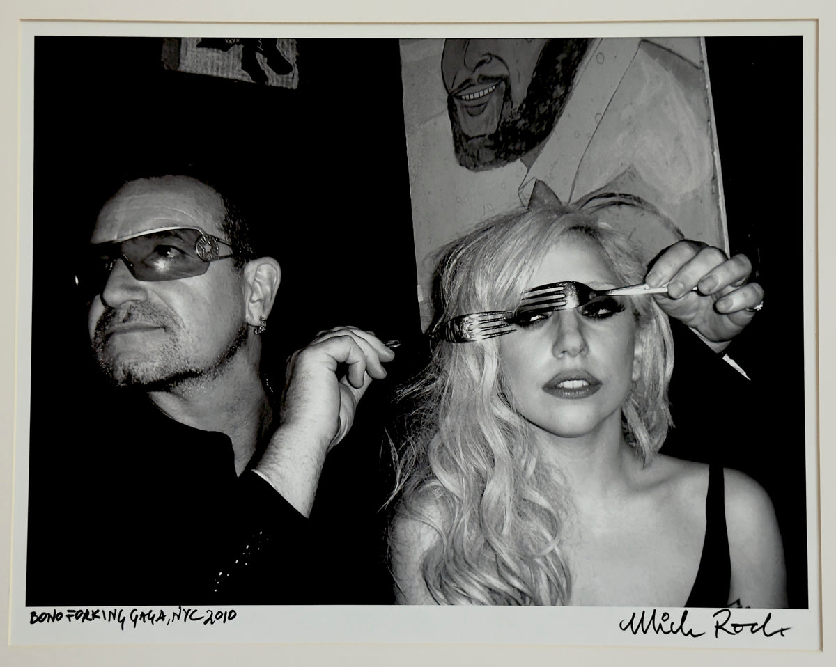 Bono and Lady Gaga at a New York club, 2010 — Lady Gaga peering through a fork as Bono looks on — photograph by Mick Rock titled ‘Bono Forking Gaga’, a signed, hand-titled late print.