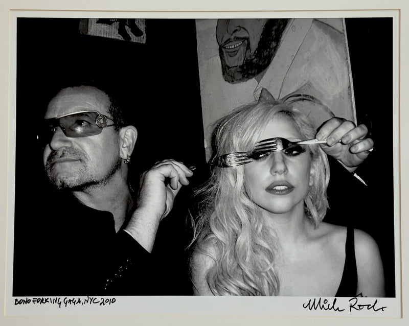 Bono and Lady Gaga at a New York club, 2010 — Lady Gaga peering through a fork as Bono looks on — photograph by Mick Rock titled ‘Bono Forking Gaga’, a signed, hand-titled late print.