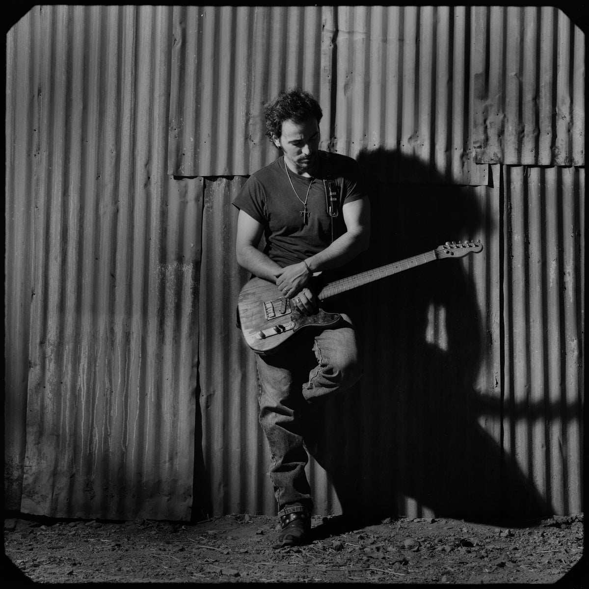 Bruce Springsteen stands with an electric guitar against a corrugated wall, its vertical lines framing his figure. Photograph by Timothy White, Malibu, California, 1991.