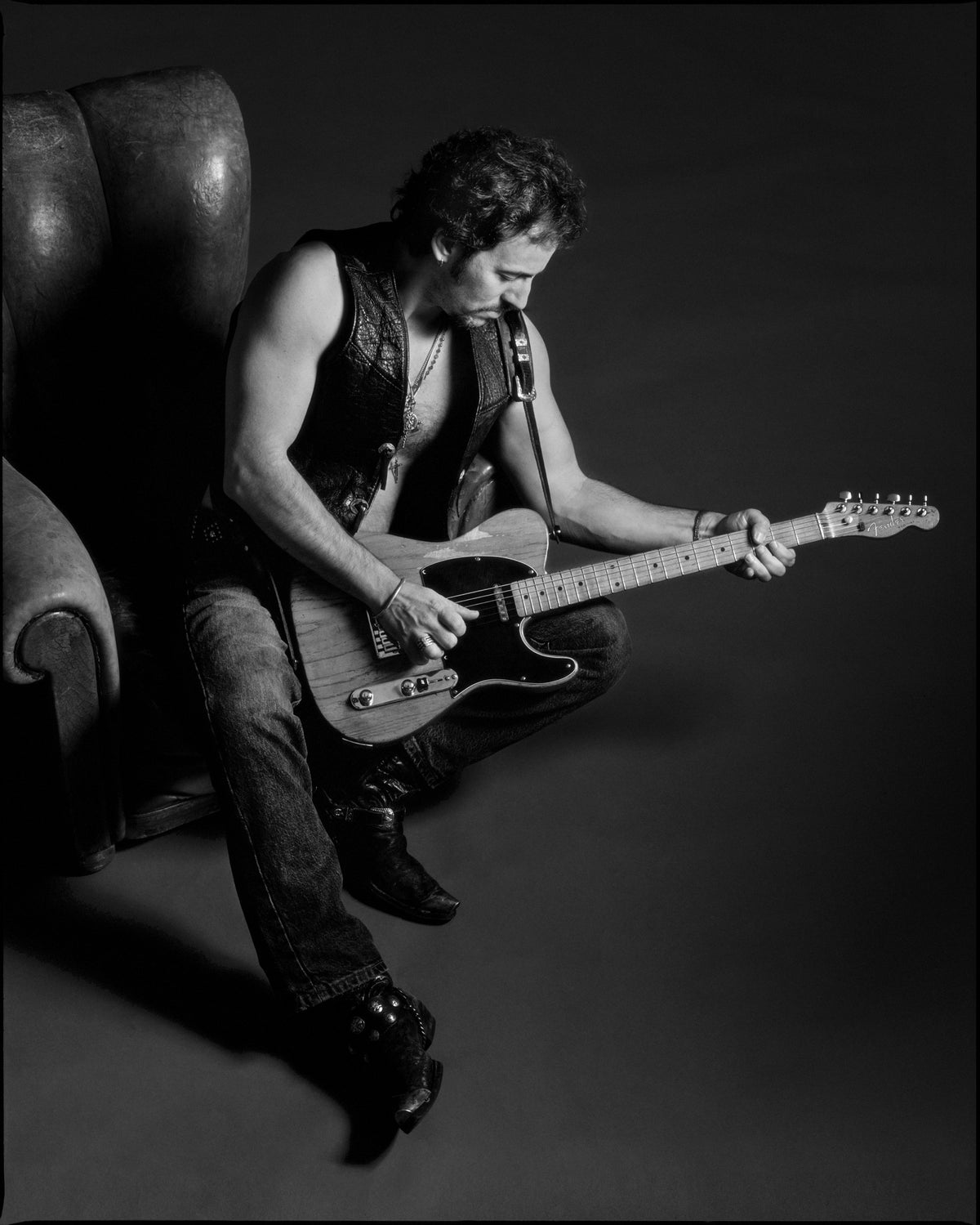 Bruce Springsteen sits at the edge of a chair, leaning over an electric guitar in a dark studio setting. Photograph by Timothy White, Malibu, California, 1991.