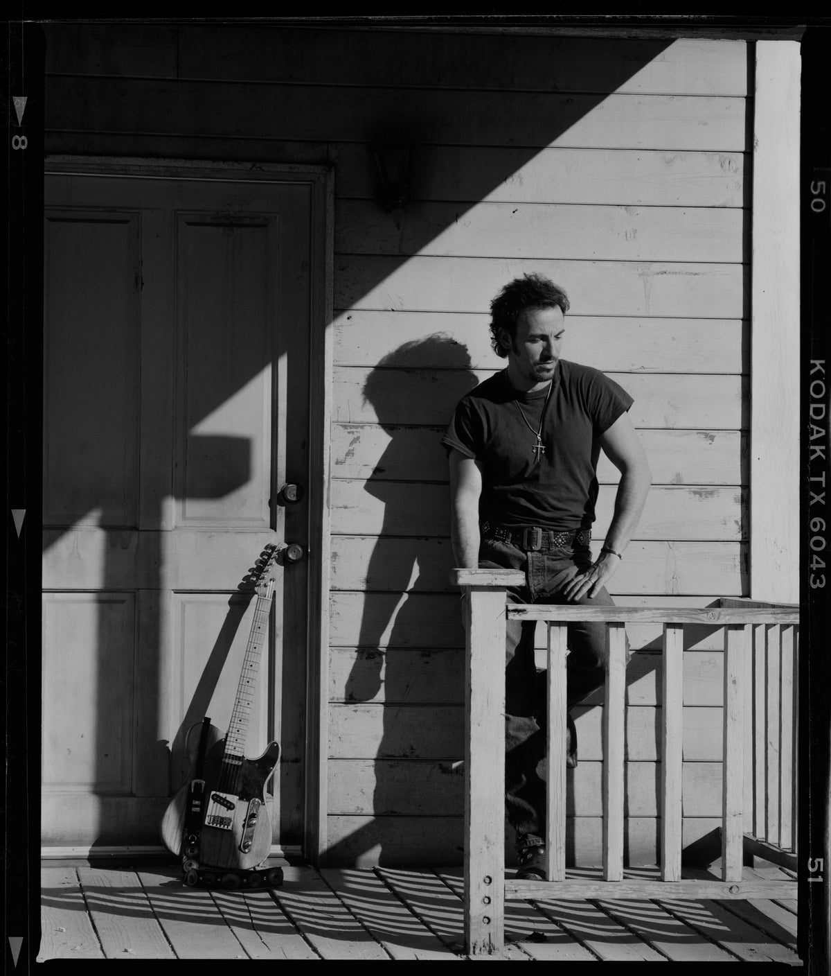 Bruce Springsteen stands on a wooden porch with his guitar, strong sunlight cutting across the wall behind him. Photograph by Timothy White, Malibu, California, 1991.