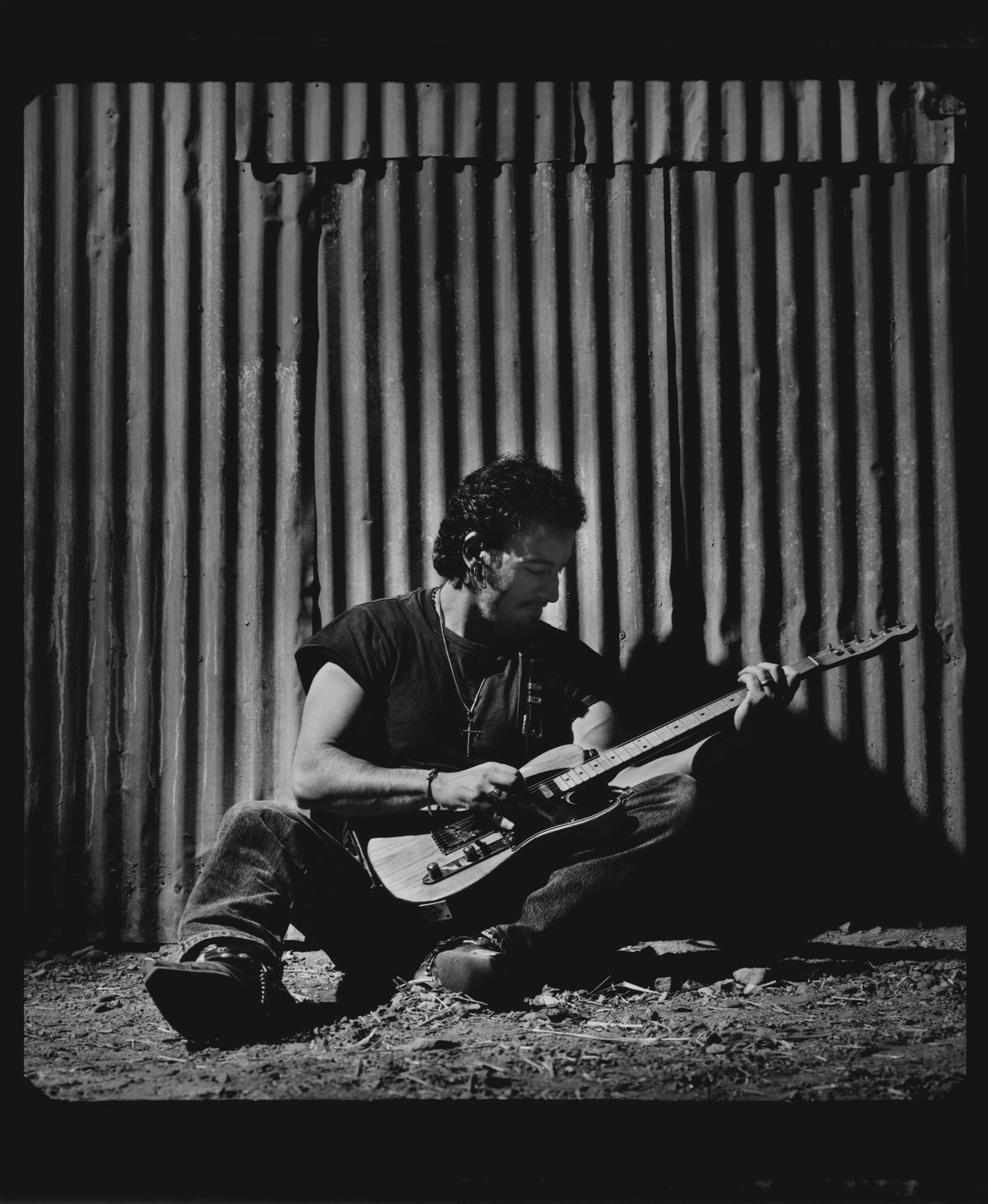 Bruce Springsteen sits on the ground with an electric guitar against a corrugated wall. Photograph by Timothy White, Malibu, California, 1991.