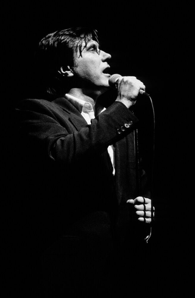 Bryan Ferry singing in a dark suit against black, in black and white, 1990, photographed by Lynn Goldsmith.