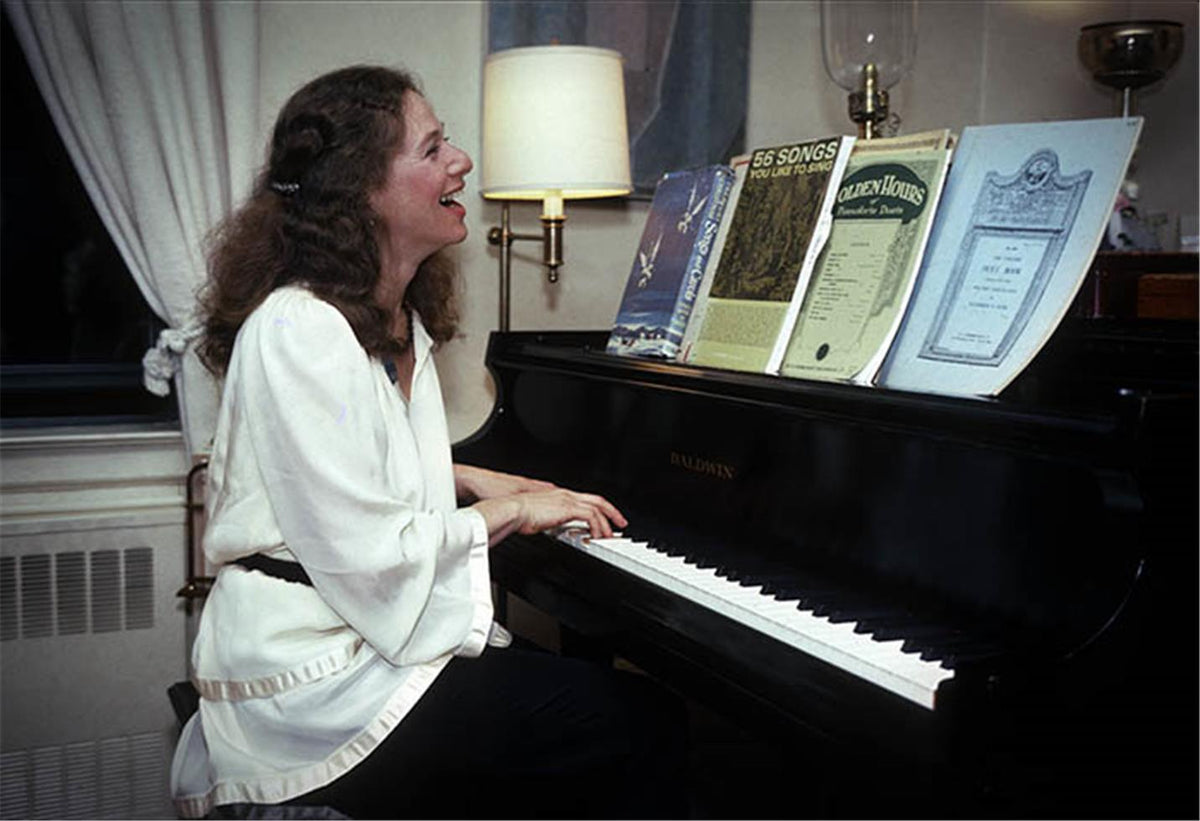 Carole King laughing at the piano in a warm interior with framed records behind her, photographed by Charlyn Zlotnik.