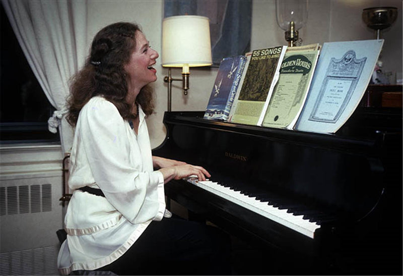 Carole King laughing at the piano in a warm interior with framed records behind her, photographed by Charlyn Zlotnik.