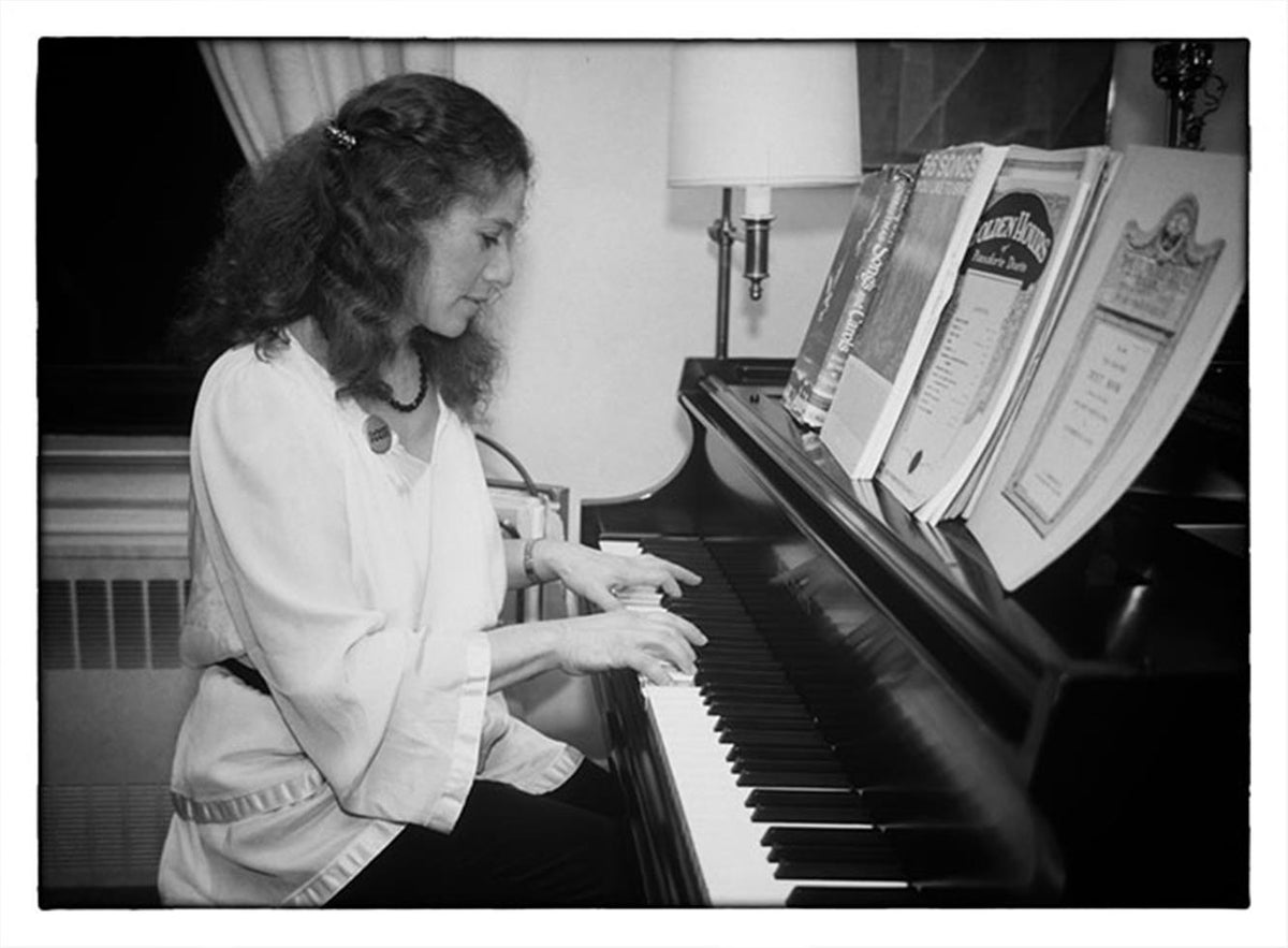 Carole King in profile playing a piano indoors with framed awards behind her, photographed by Charlyn Zlotnik.
