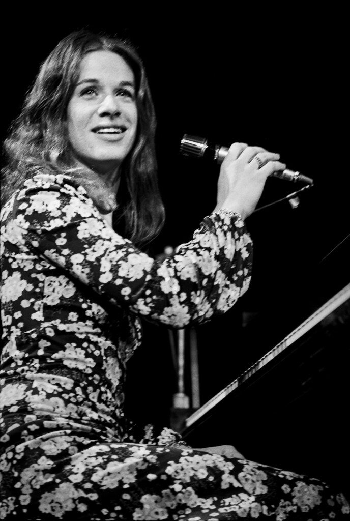 Carole King smiling at the piano with a microphone, in black and white, 1972, photographed by Lynn Goldsmith.