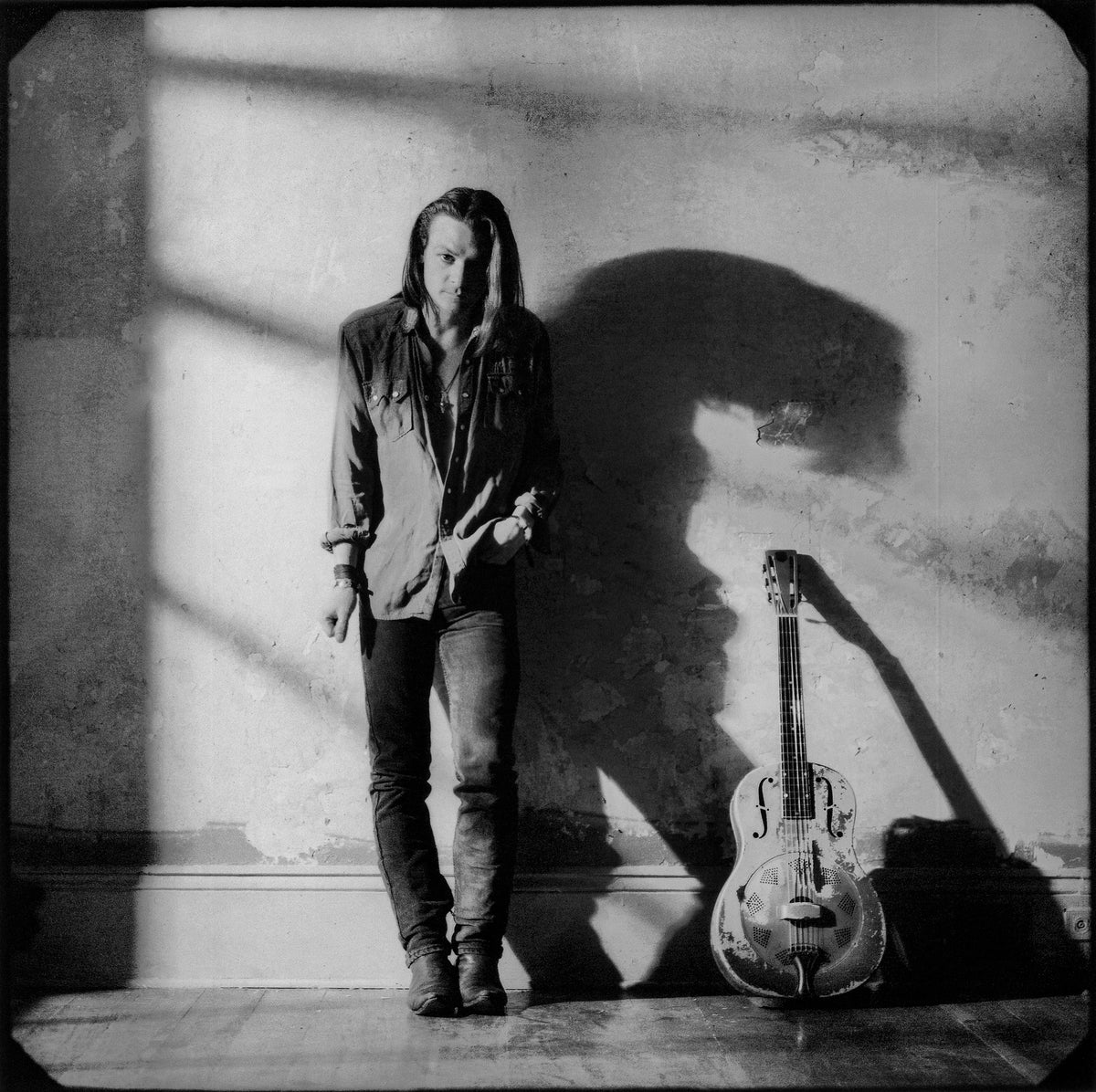 Chris Whitley stands beside his guitar as strong light casts his shadow across a bare wall. Photograph by Timothy White, New Orleans, Louisiana, 1991.