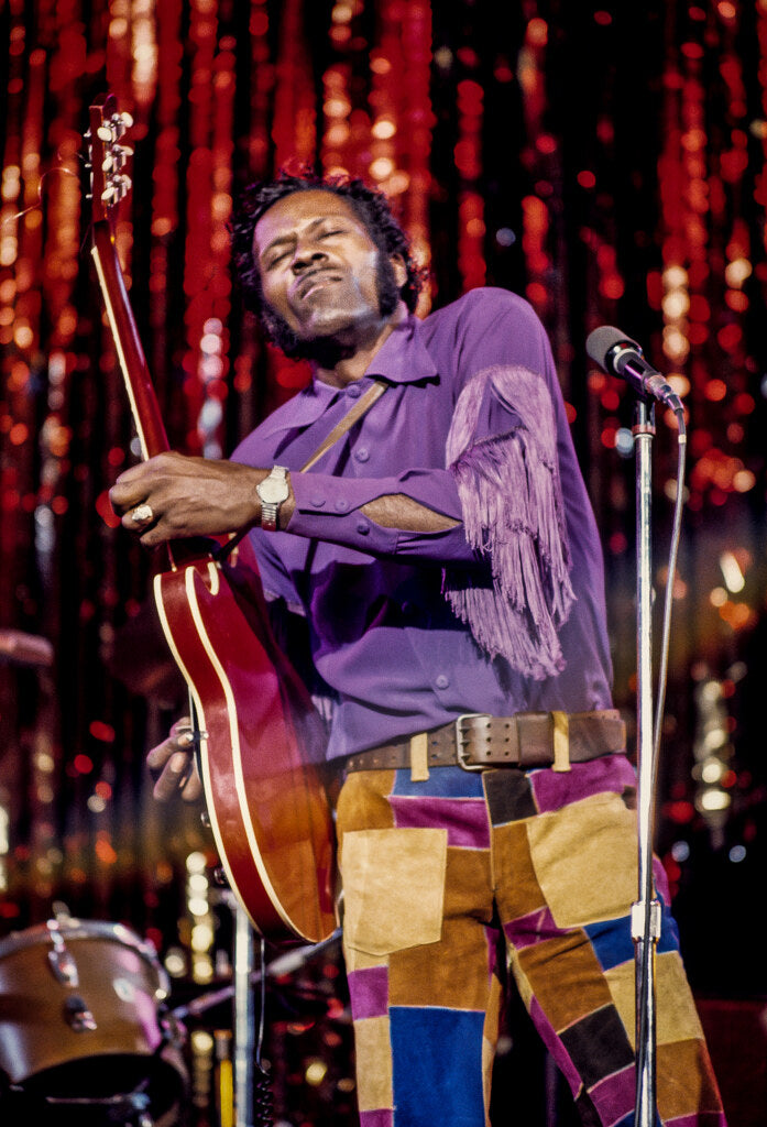 Chuck Berry with his guitar in a purple fringed shirt before a red tinsel curtain, 1974, photographed by Lynn Goldsmith.