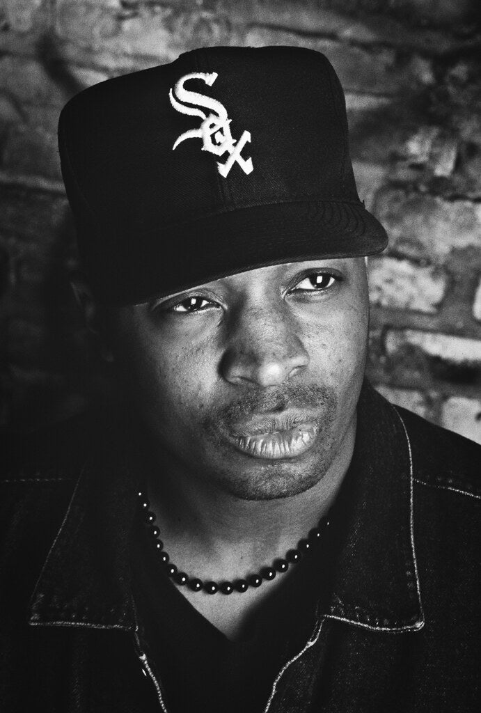 Close portrait of Chuck D in a baseball cap against brick, in black and white, 1991, photographed by Lynn Goldsmith.