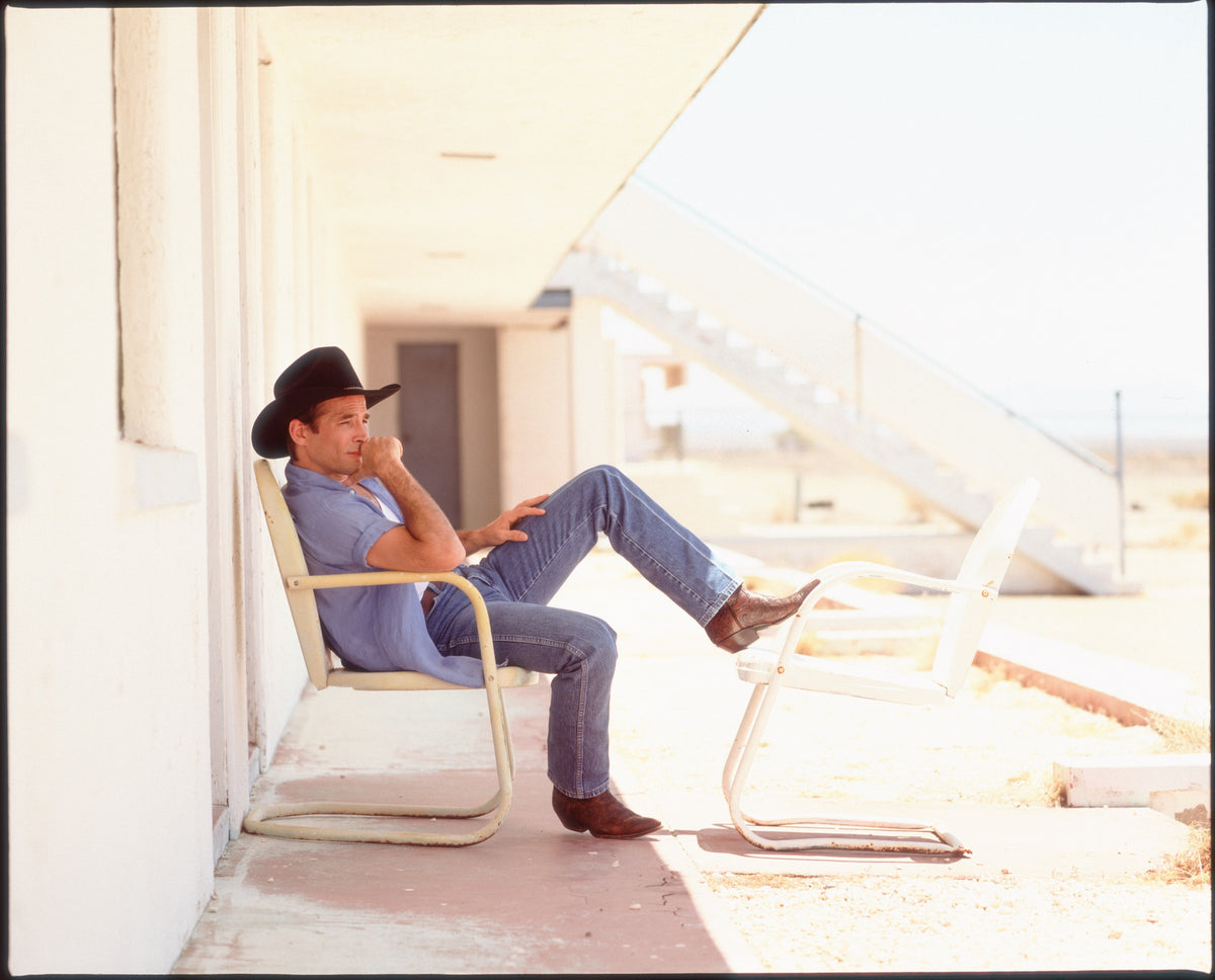 Clint Black sits on a chair outside a pale building, the desert light washing across the scene. Photograph by Timothy White, Amboy, California, 1997.