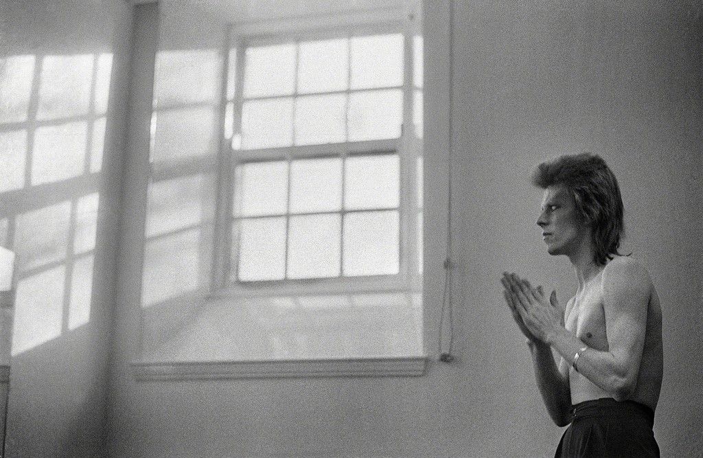 Black-and-white photograph of David Bowie standing by a tall window, Scotland, 1973, photographed by Mick Rock.