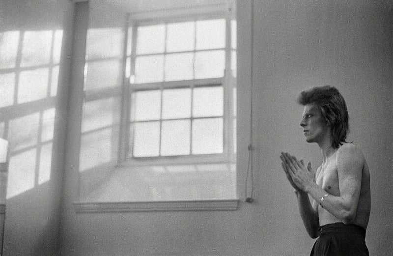 Black-and-white photograph of David Bowie standing by a tall window, Scotland, 1973, photographed by Mick Rock.