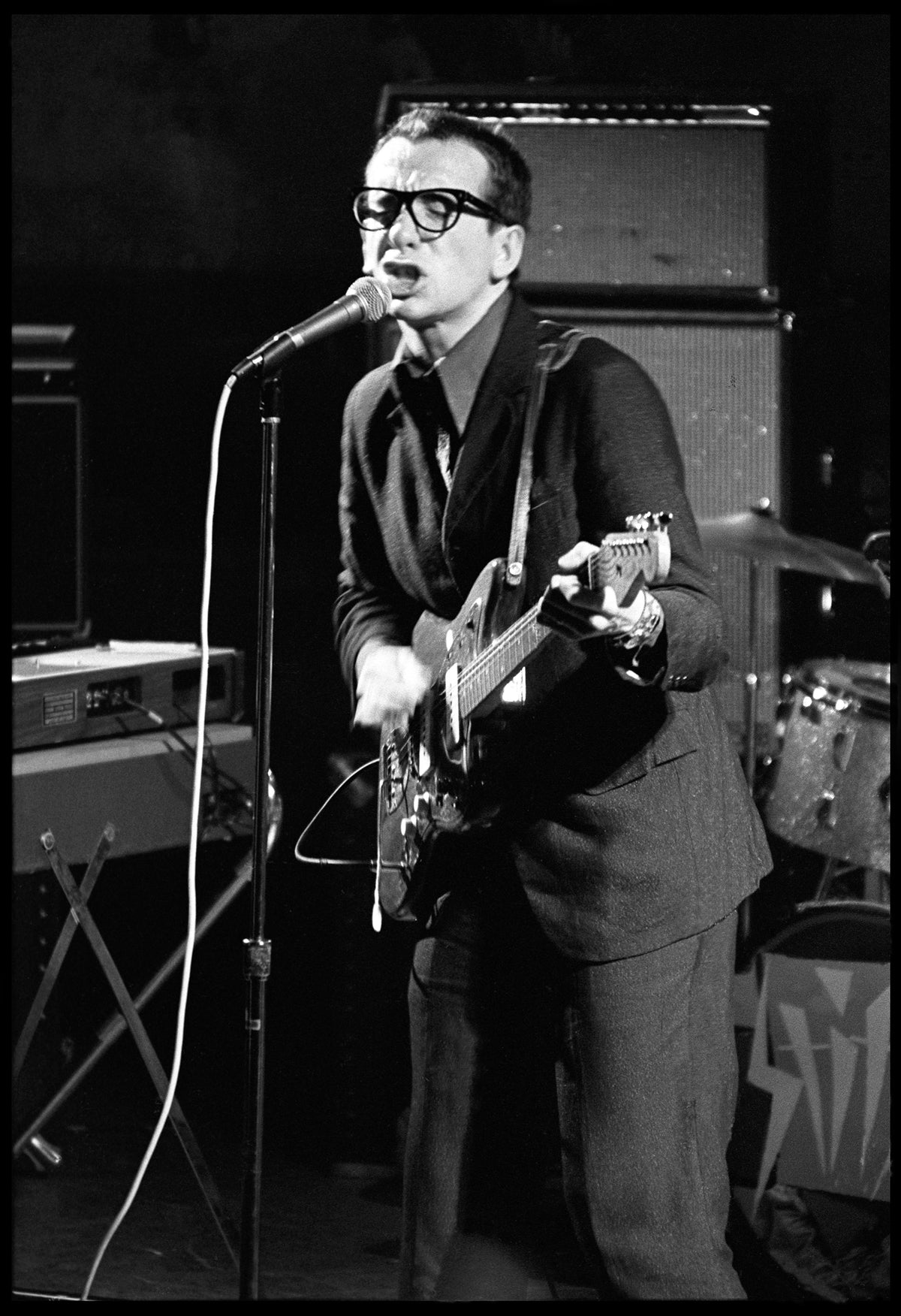 Elvis Costello singing into a microphone and playing guitar on stage at Birmingham Town Hall, 1977, photographed by David Corio.