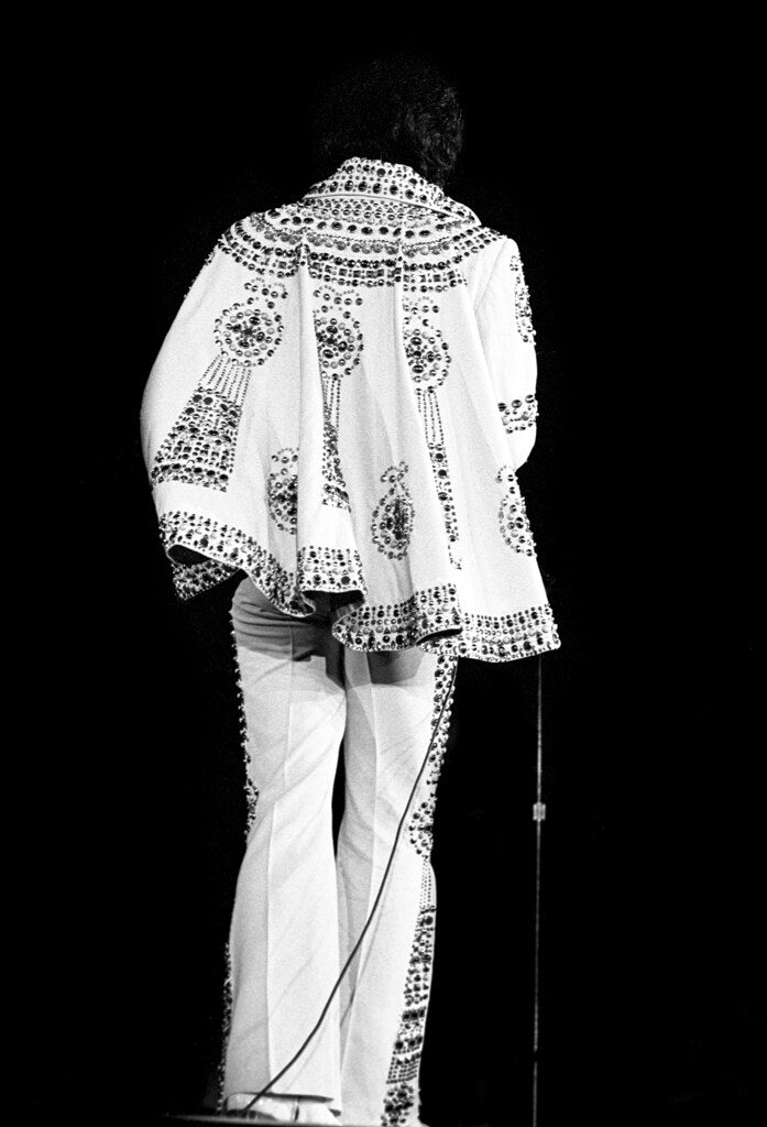 Elvis Presley seen from behind on stage in a white embroidered jumpsuit, 1974, photographed by Lynn Goldsmith.