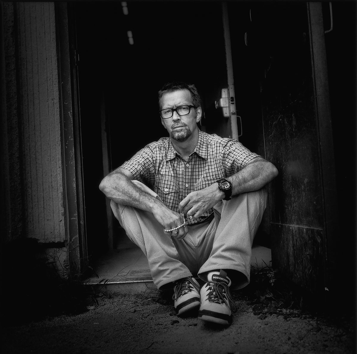 Eric Clapton sits against a dark wooden wall, wearing a beaded necklace and looking toward the camera. Photograph by Timothy White, Toronto, Canada, 1997.