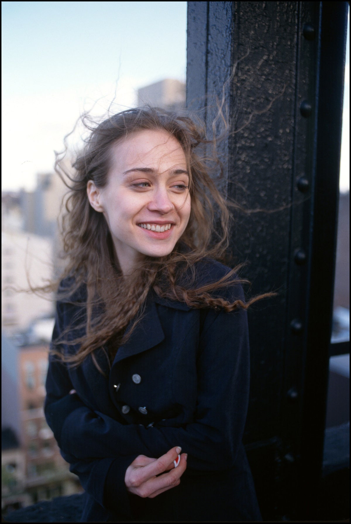 Fiona Apple smiling on a rooftop with windblown hair, wearing a dark coat, Gramercy Park, New York City, 1997, photographed in color by David Corio.