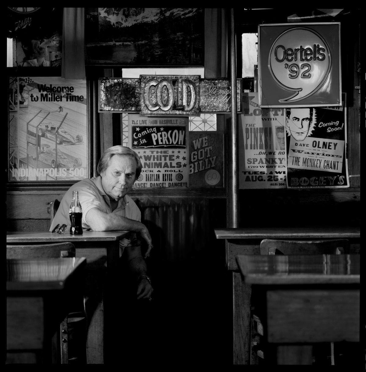 George Jones sits beneath a wall of signs in a bar, his figure illuminated against the busy interior. Photograph by Timothy White, Nashville, Tennessee, 1989.