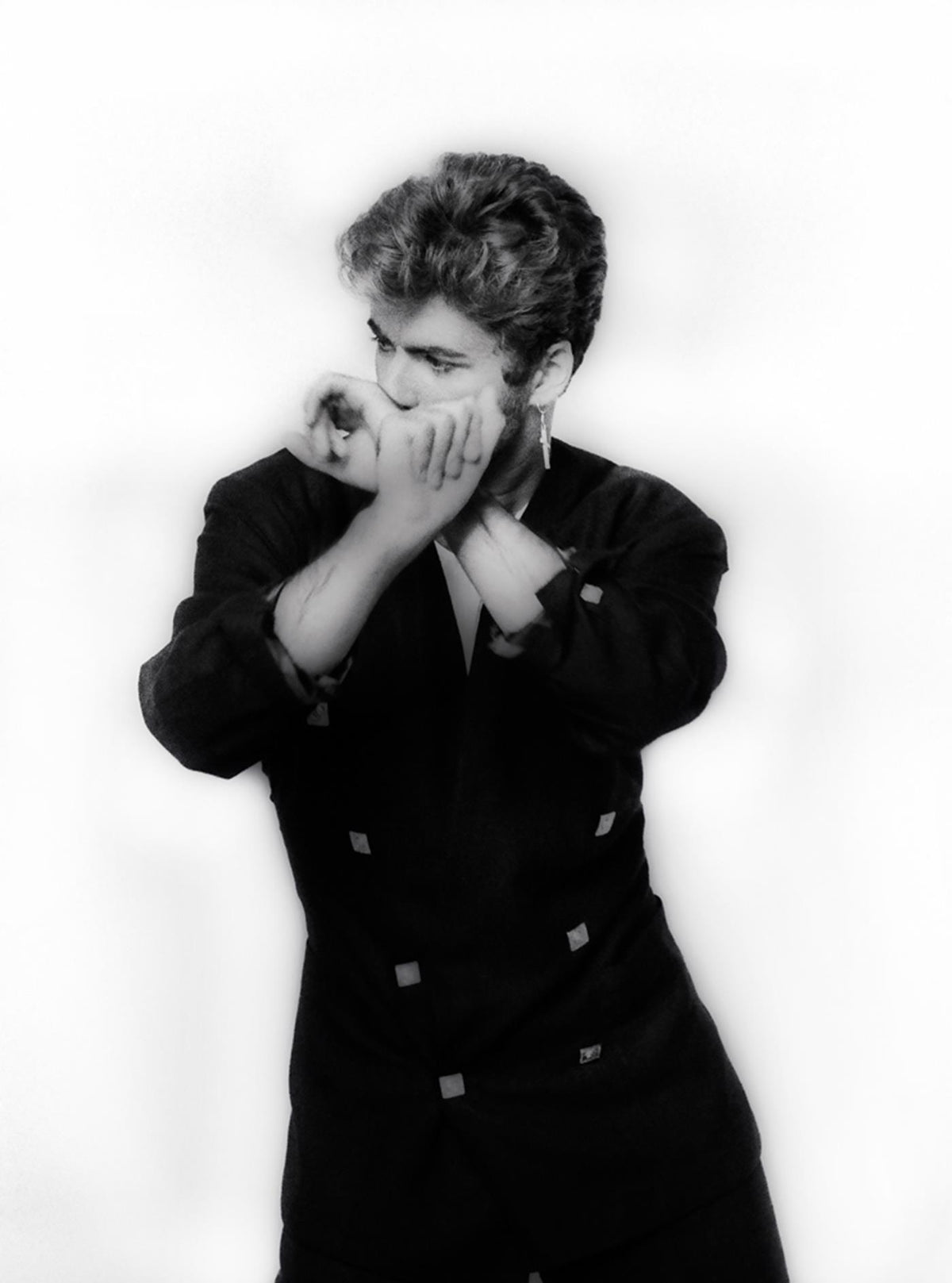 Black-and-white studio portrait of George Michael in a dark jacket, hands raised to his face, photographed by Martyn Goddard, 1987.