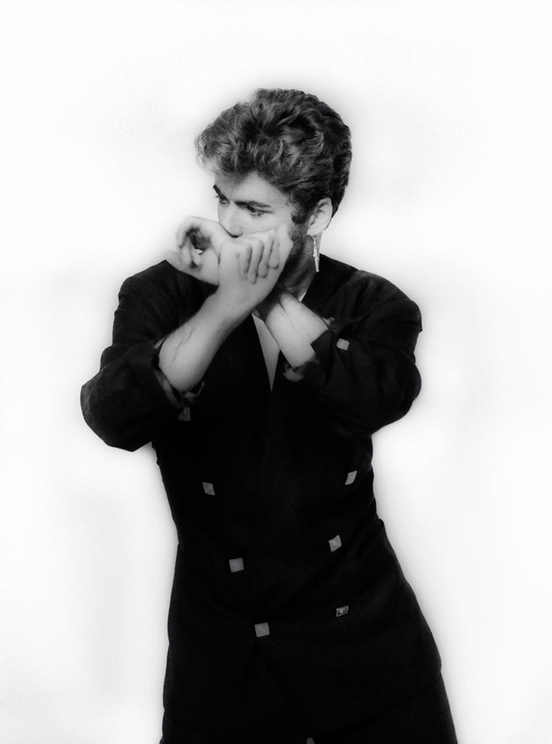 Black-and-white studio portrait of George Michael in a dark jacket, hands raised to his face, photographed by Martyn Goddard, 1987.