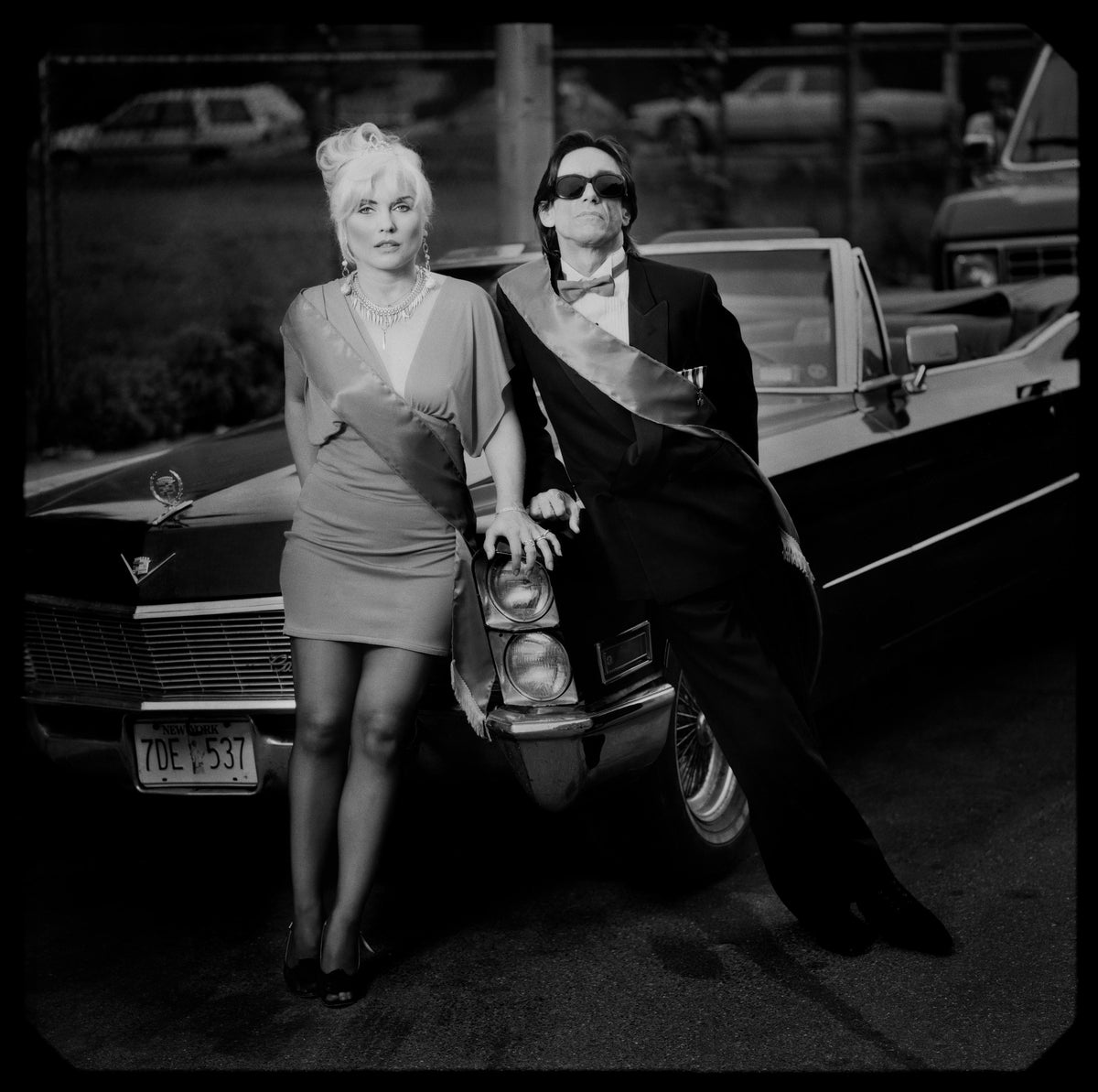 Iggy Pop and Debbie Harry pose beside a car, leaning together in an urban setting. Photograph by Timothy White, Jersey City, New Jersey, 1990.