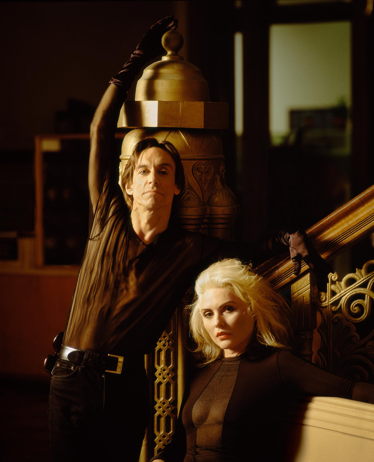 Iggy Pop raises an arm beside Debbie Harry on an interior staircase, their figures framed by warm wood tones. Photograph by Timothy White, Jersey City, New Jersey, 1990.