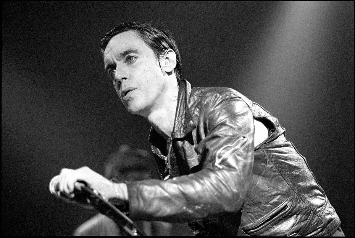 Iggy Pop in a leather jacket leaning forward with a microphone on stage at The Rainbow, London, 1981, photographed by David Corio.