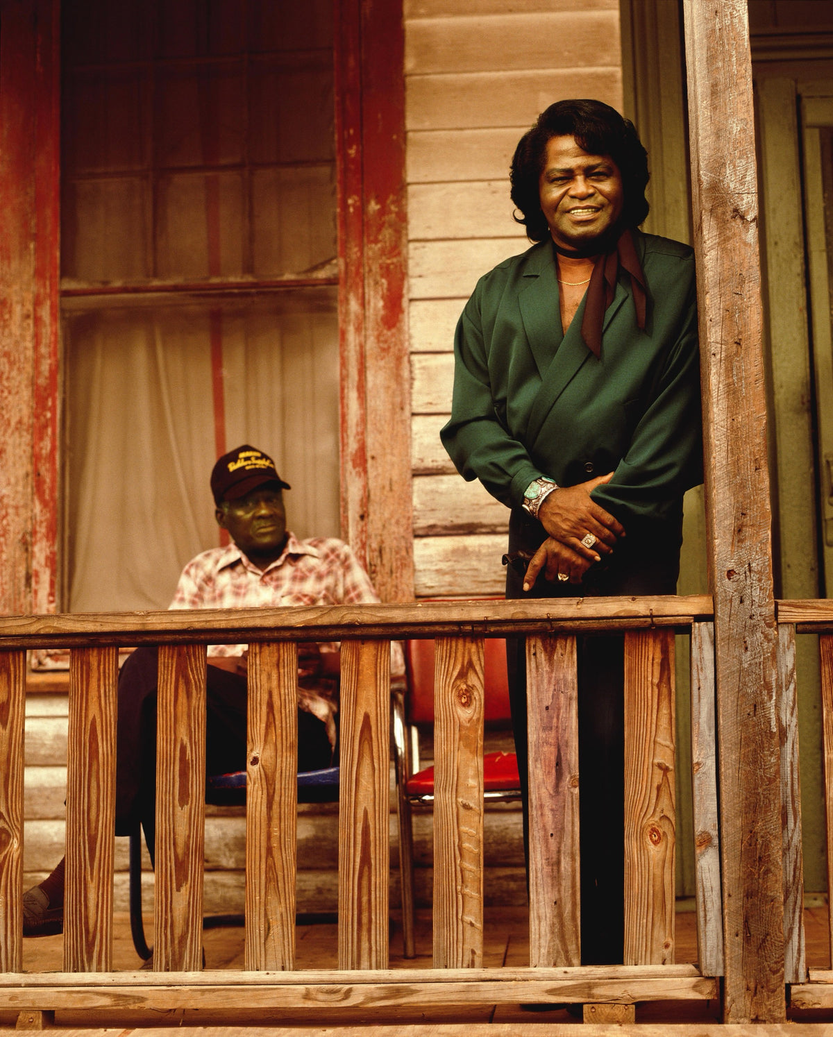 James Brown stands smiling on a wooden porch, with a second figure seated behind the railing. Photograph by Timothy White, Augusta, Georgia, 1991.