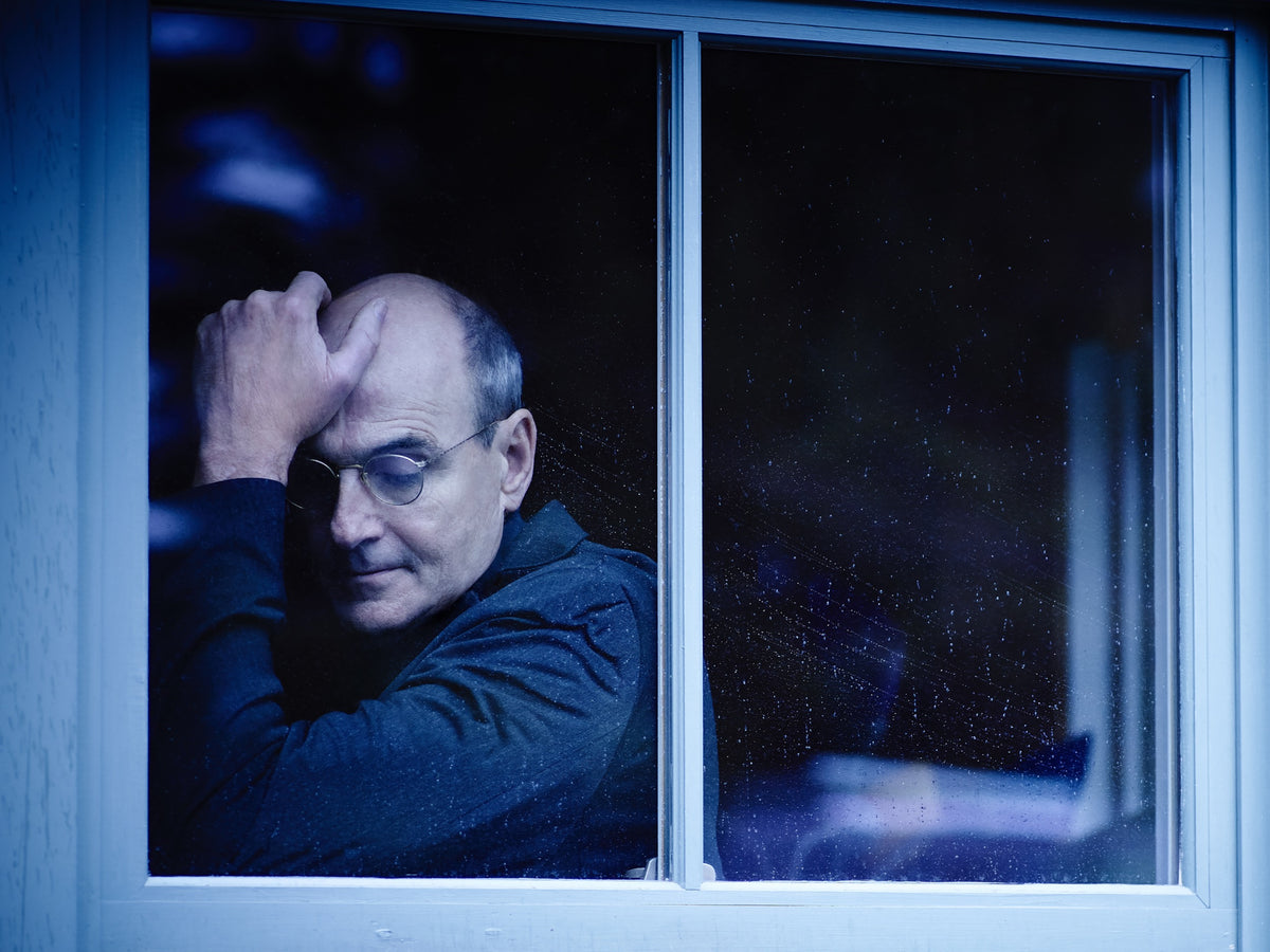 James Taylor is seen through a blue window frame, resting his hand against his head. Photograph by Timothy White, Stockbridge, Massachusetts, 2014.