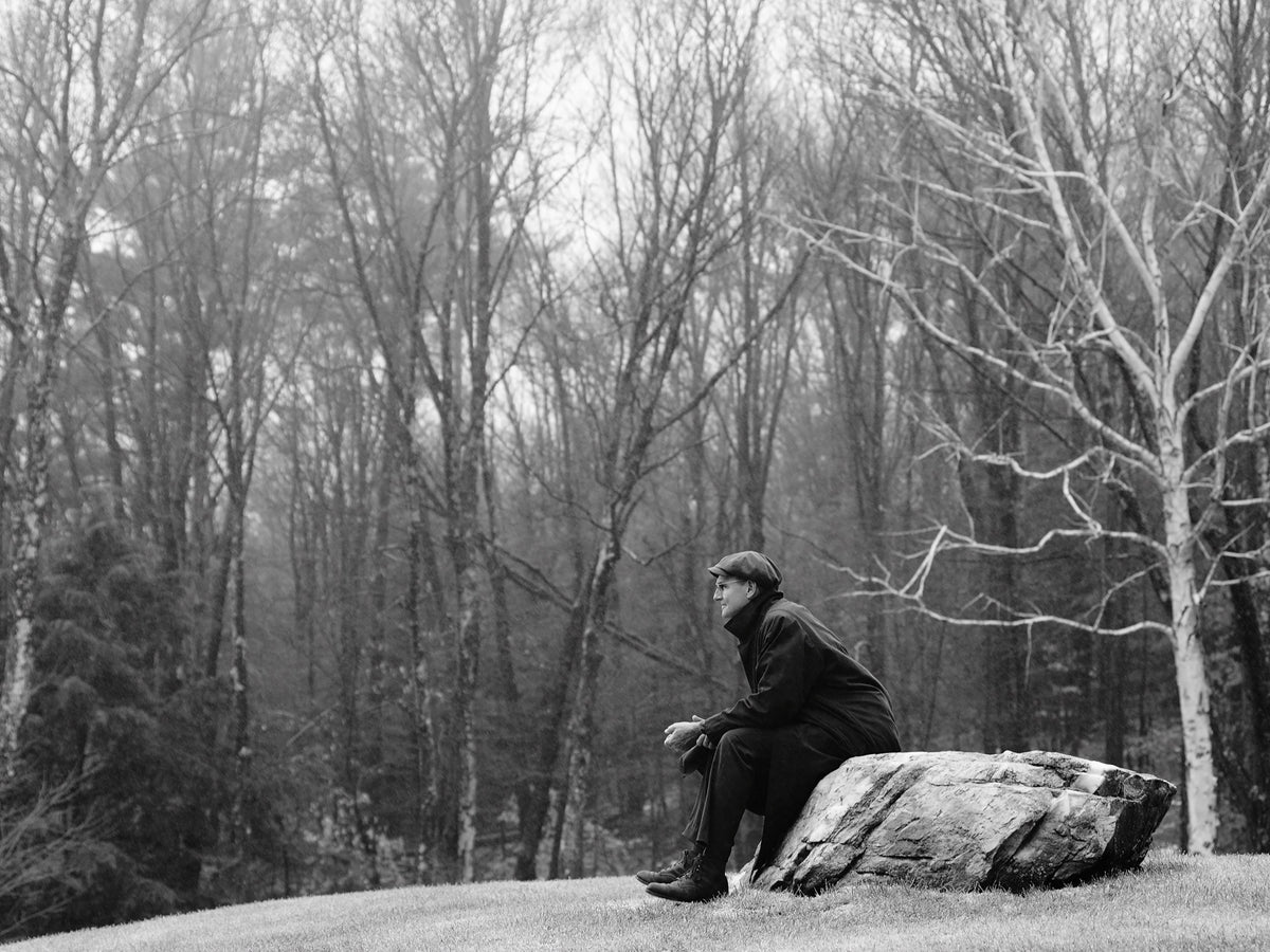 James Taylor sits on a large rock in a winter landscape, with bare trees filling the background. Photograph by Timothy White, Stockbridge, Massachusetts, 2014.