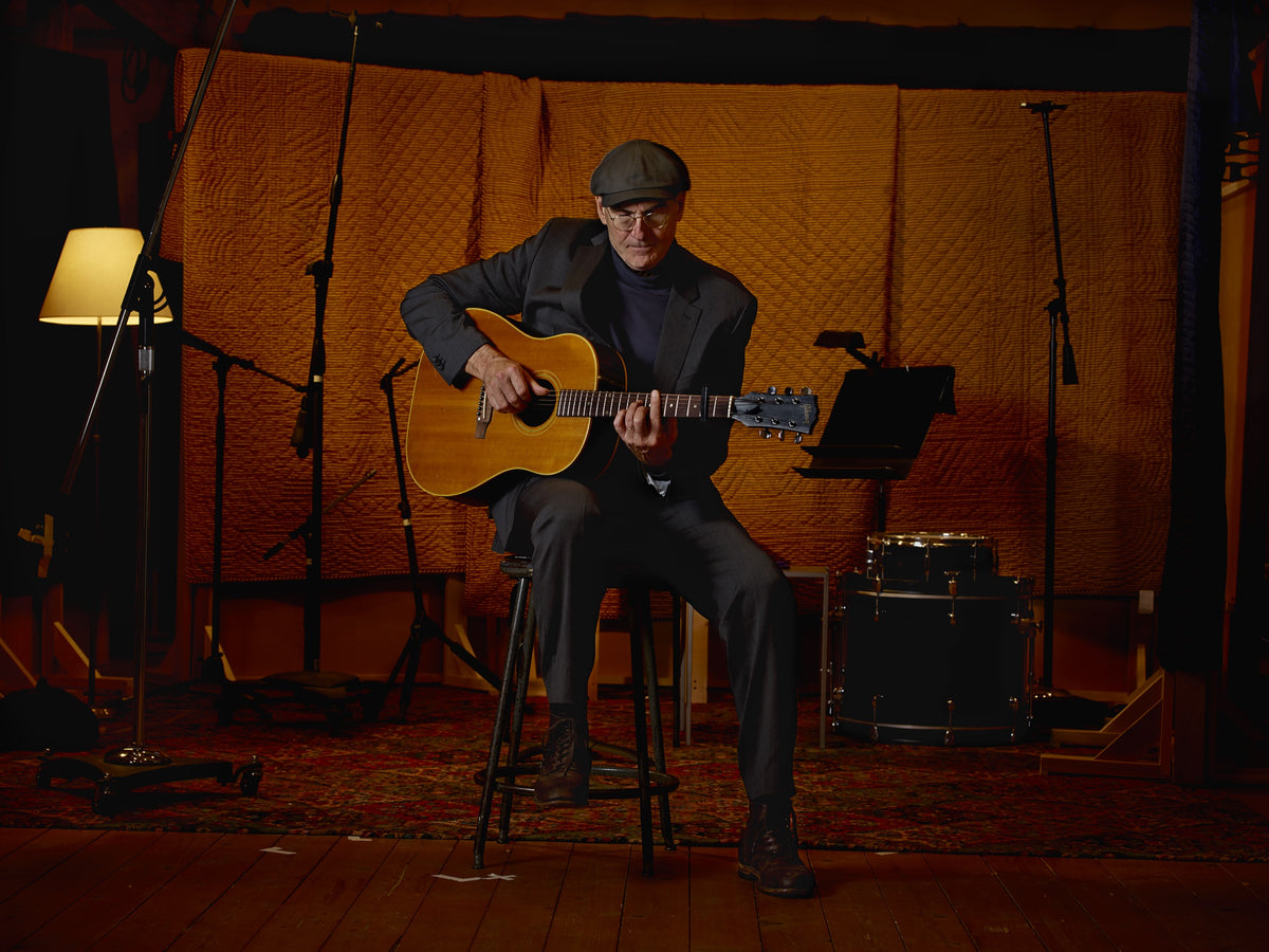 James Taylor sits on a stool playing an acoustic guitar in a warmly lit recording space. Photograph by Timothy White, Stockbridge, Massachusetts, 2014.