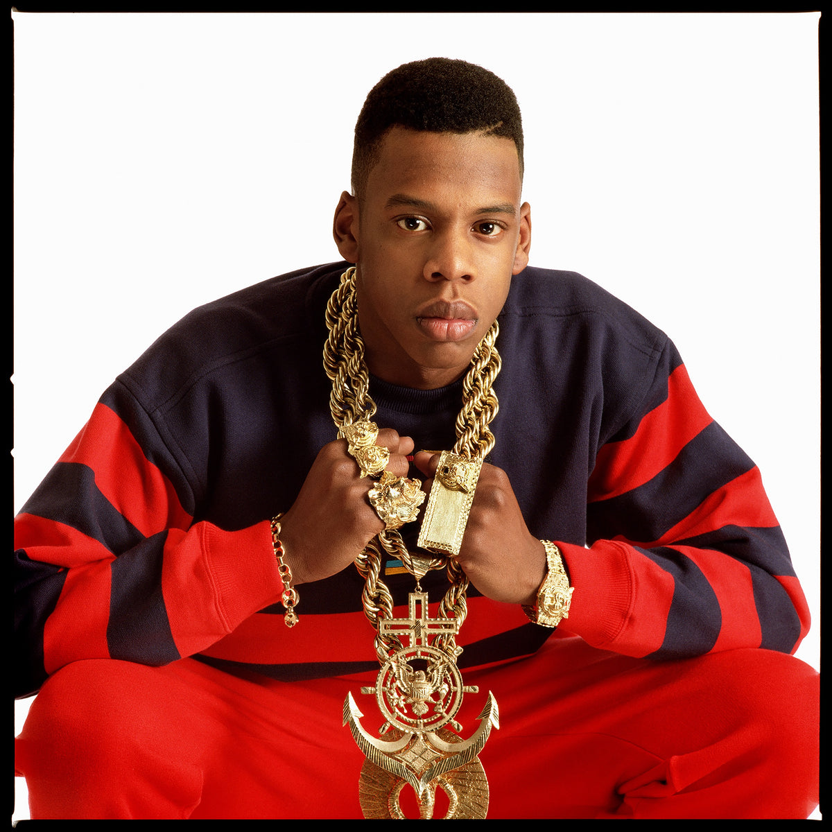 Jay-Z faces the camera in a red and black sweater, holding layers of gold chains at his chest. Photograph by Timothy White, New York City, 1988.