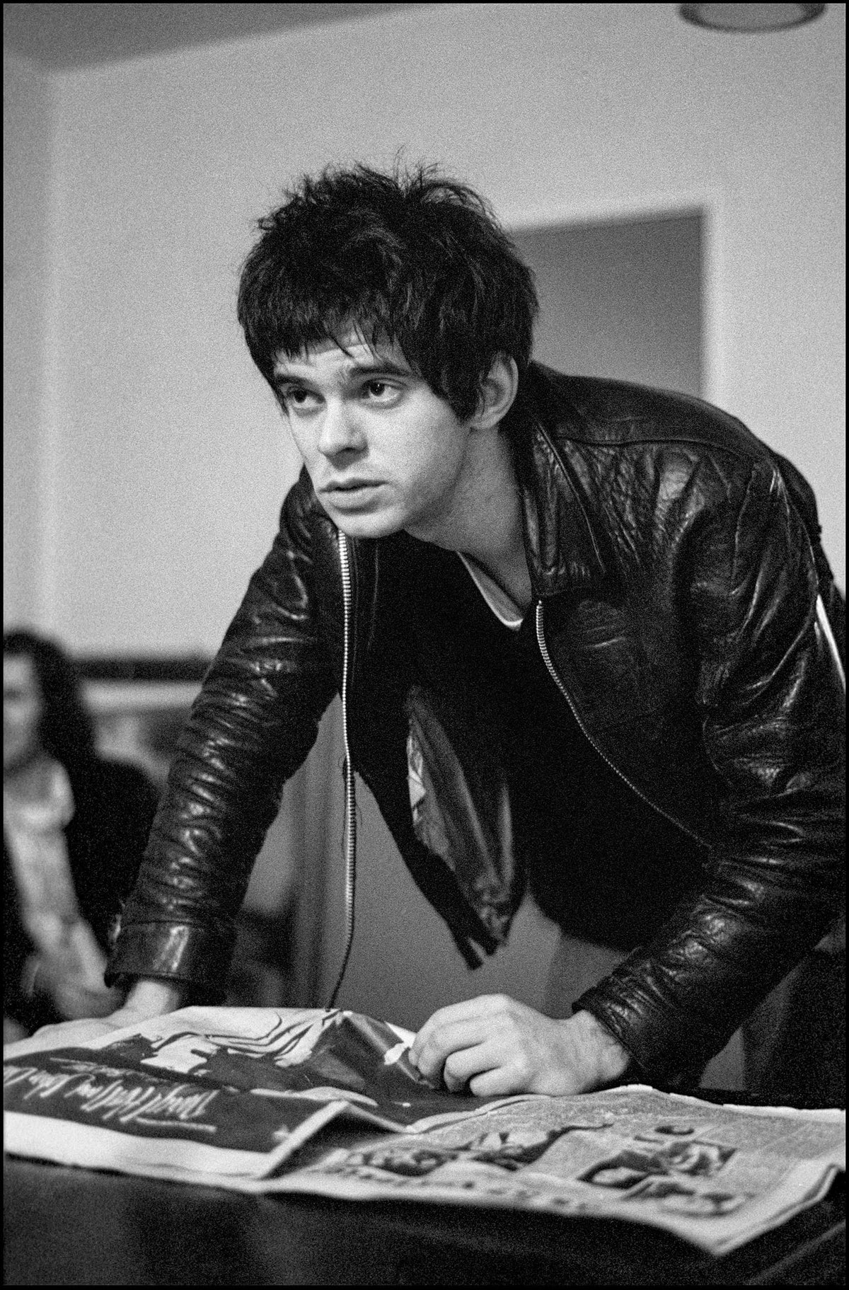 Jean-Jacques Burnel of The Stranglers in a leather jacket leaning over a copy of the NME backstage at Malvern Winter Gardens, 1977, photographed by David Corio.