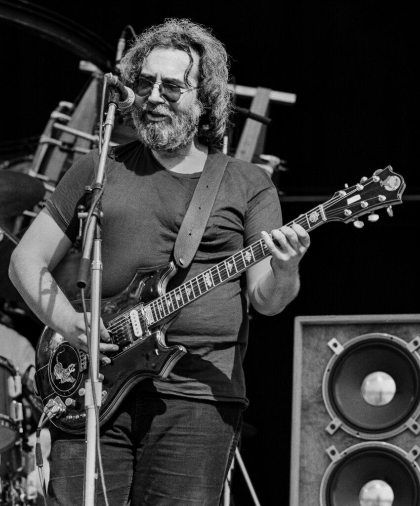 Jerry Garcia playing guitar on an outdoor stage, in black and white, 1982, photographed by Lynn Goldsmith.