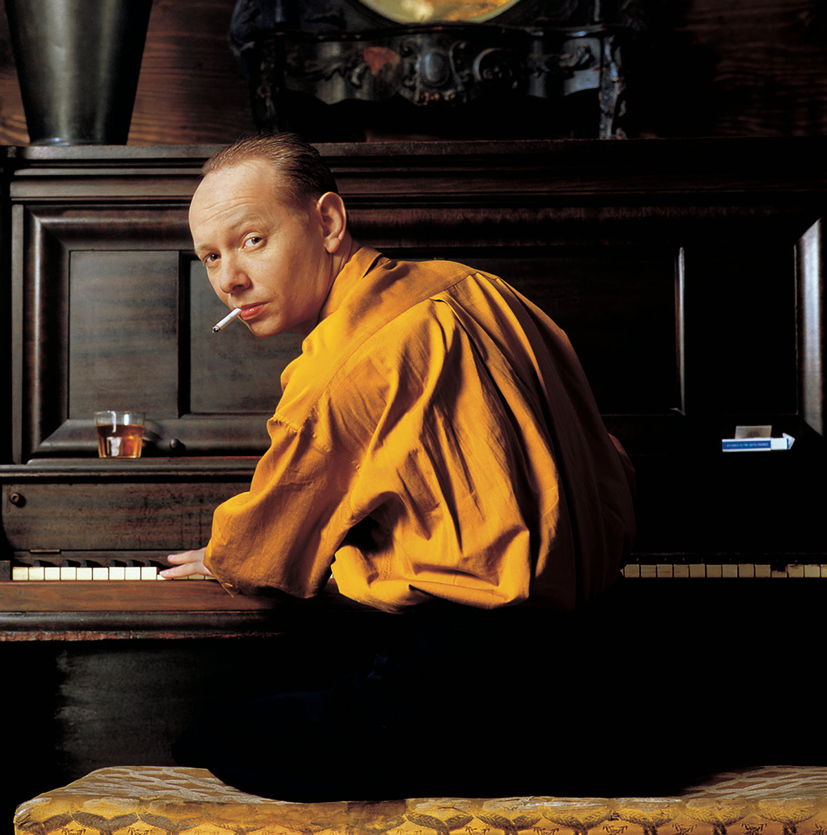 Seated at a piano, Joe Jackson turns toward the camera in a warmly lit interior. Photograph by Timothy White, New York City, 1989.