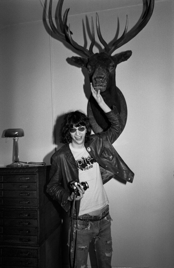 Joey Ramone in a leather jacket beneath a mounted elk head, in black and white, 1977, photographed by Lynn Goldsmith.