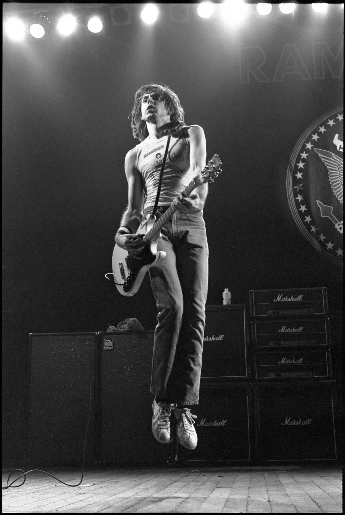 Johnny Ramone jumping in the air with his guitar in front of Marshall amplifiers and stage lights, Hammersmith Odeon, London, 1980, photographed by David Corio.