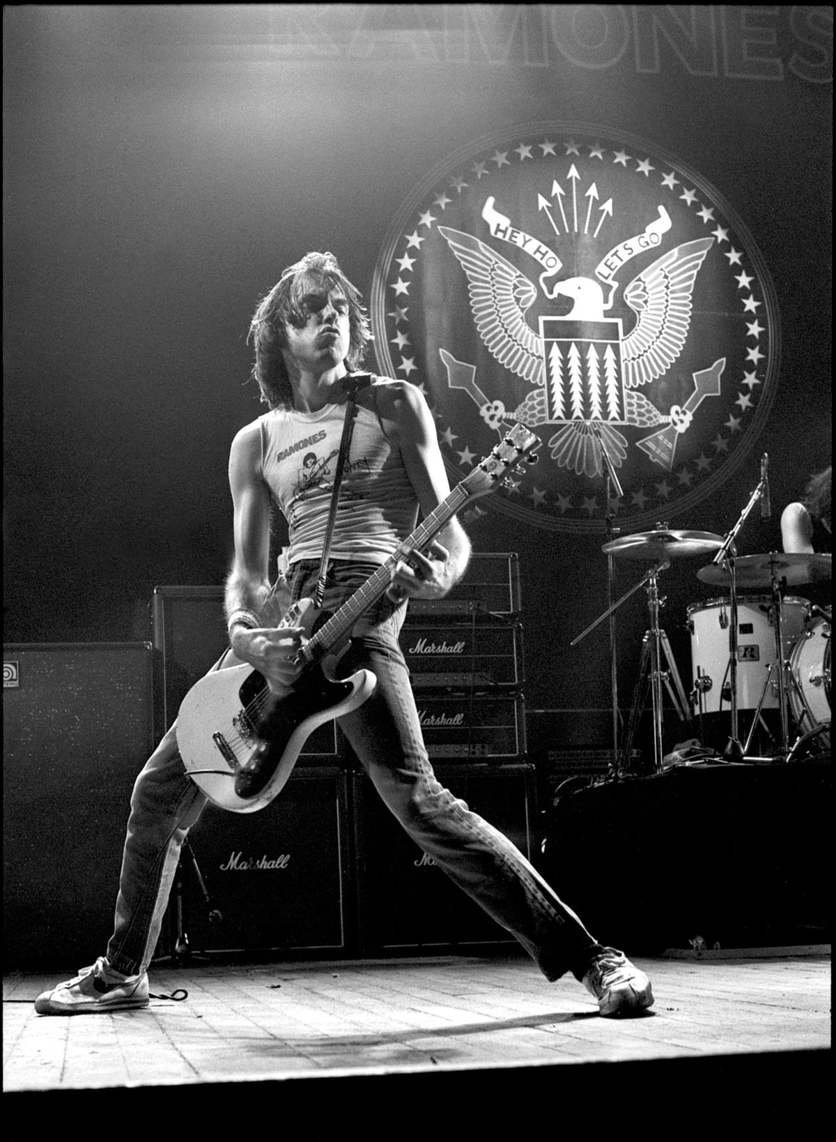 Johnny Ramone playing guitar with legs spread wide in front of the Ramones eagle seal backdrop, Hammersmith Odeon, London, 1980, photographed by David Corio.